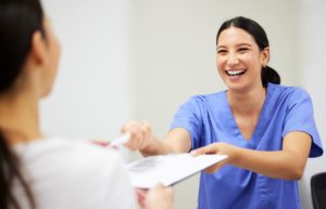 Woman with dark hair in blue scrubs smiling and handing a patient forms to fill out